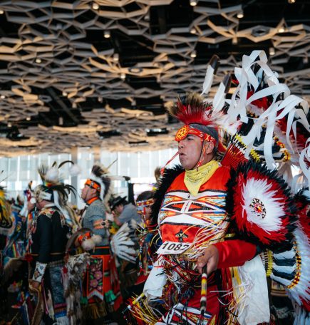 Indigenous person dressed in traditional head dress, dancing with others at Manito Ahbee Festival pow wow.