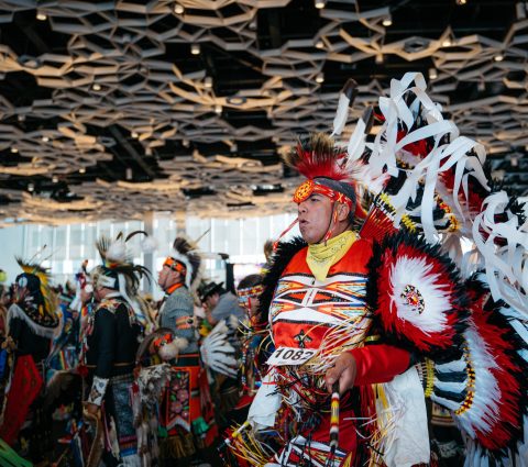 Indigenous person dressed in traditional head dress, dancing with others at Manito Ahbee Festival pow wow.