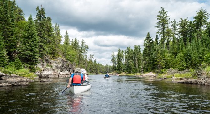 Two canoes travel down a river with tree-lined shores.